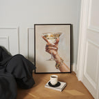 a framed painting of a hand holding a martini glass, with a cup of coffee and a book on a wooden floor in front of it.