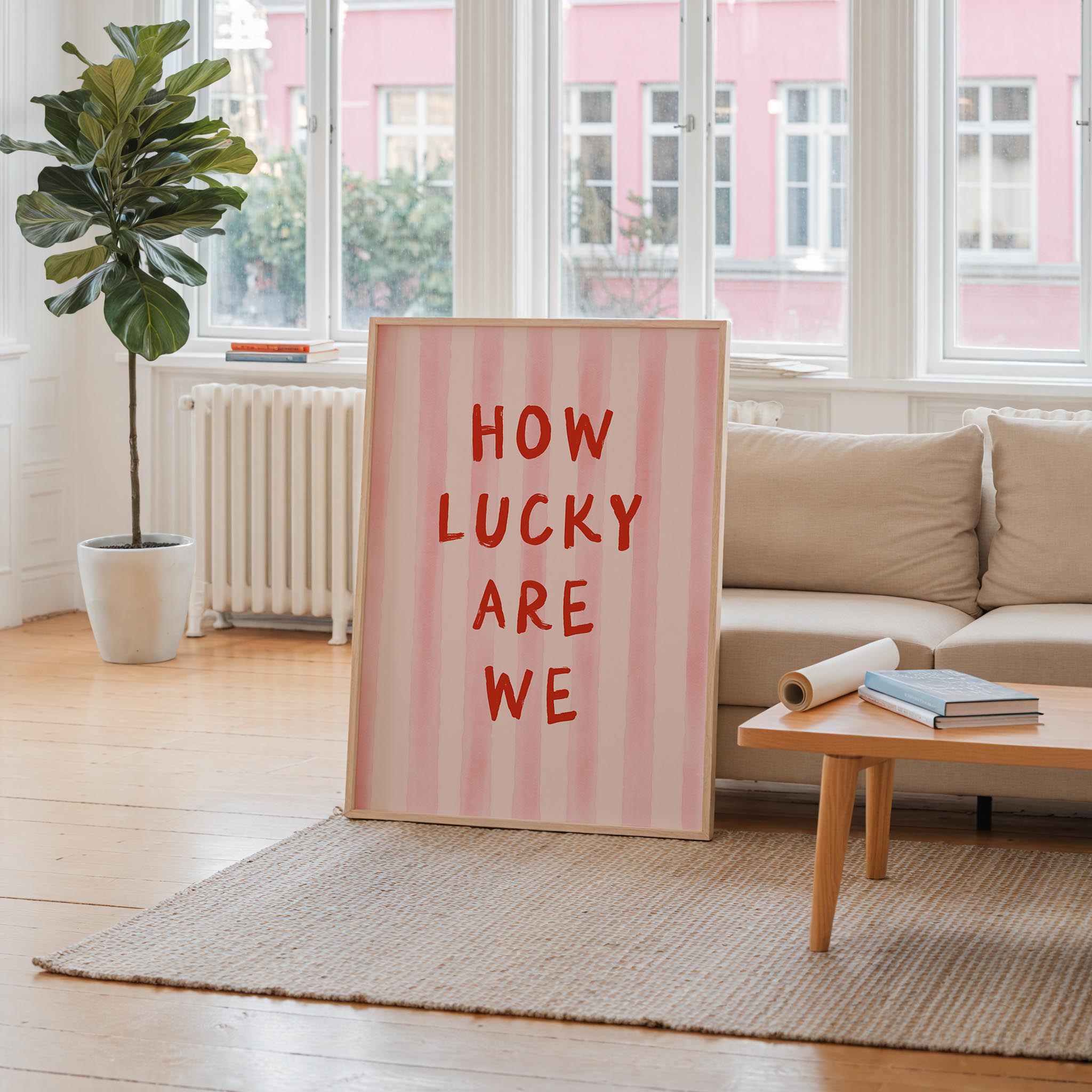 a large pink sign with the text "HOW LUCKY ARE WE" in bold red letters, placed in a living room setting with a couch, a coffee table, and a potted plant.