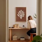 A woman is sitting at a wooden desk, surrounded by various objects, including a framed plant-like artwork, books, and a stack of books.