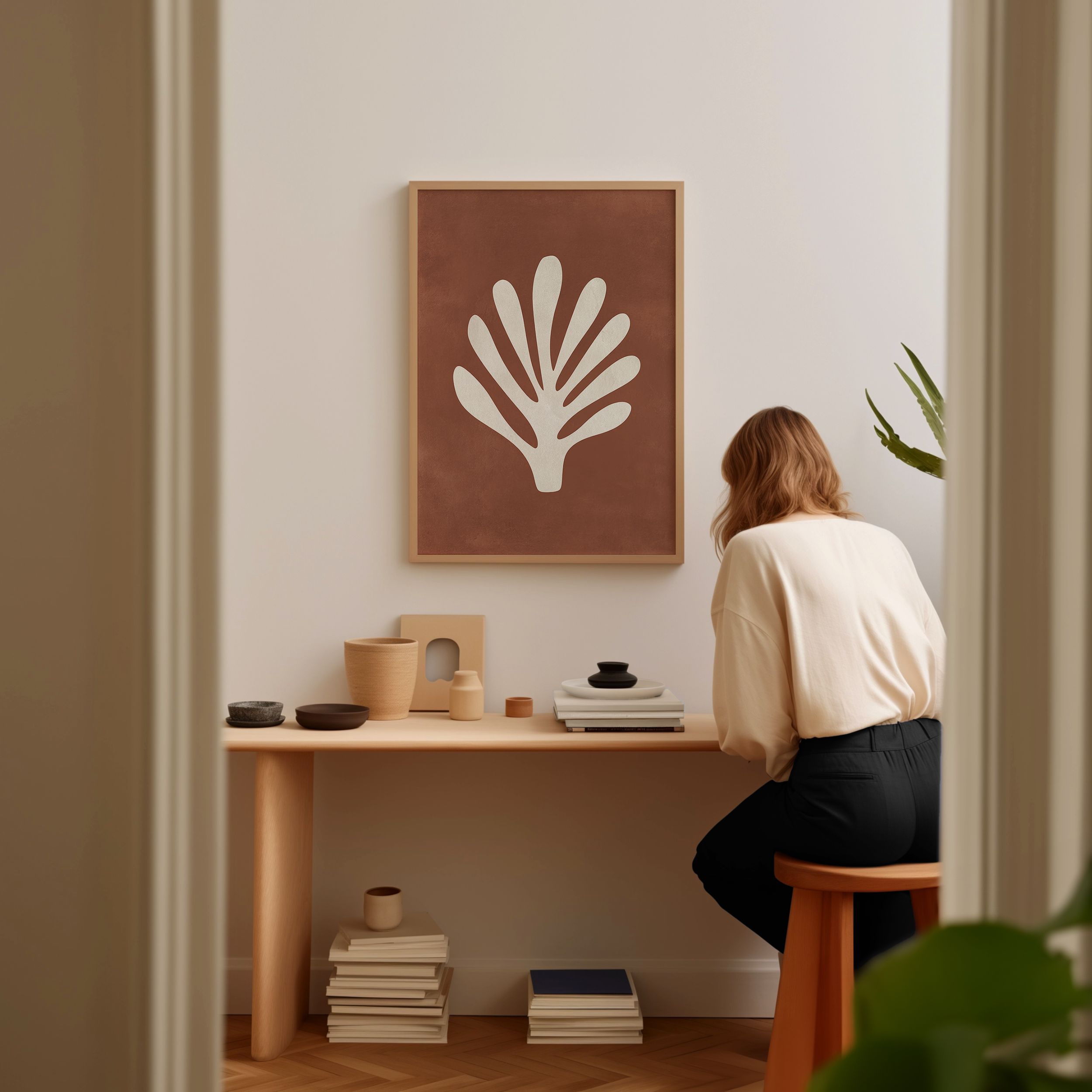 A woman is sitting at a wooden desk, surrounded by various objects, including a framed plant-like artwork, books, and a stack of books.