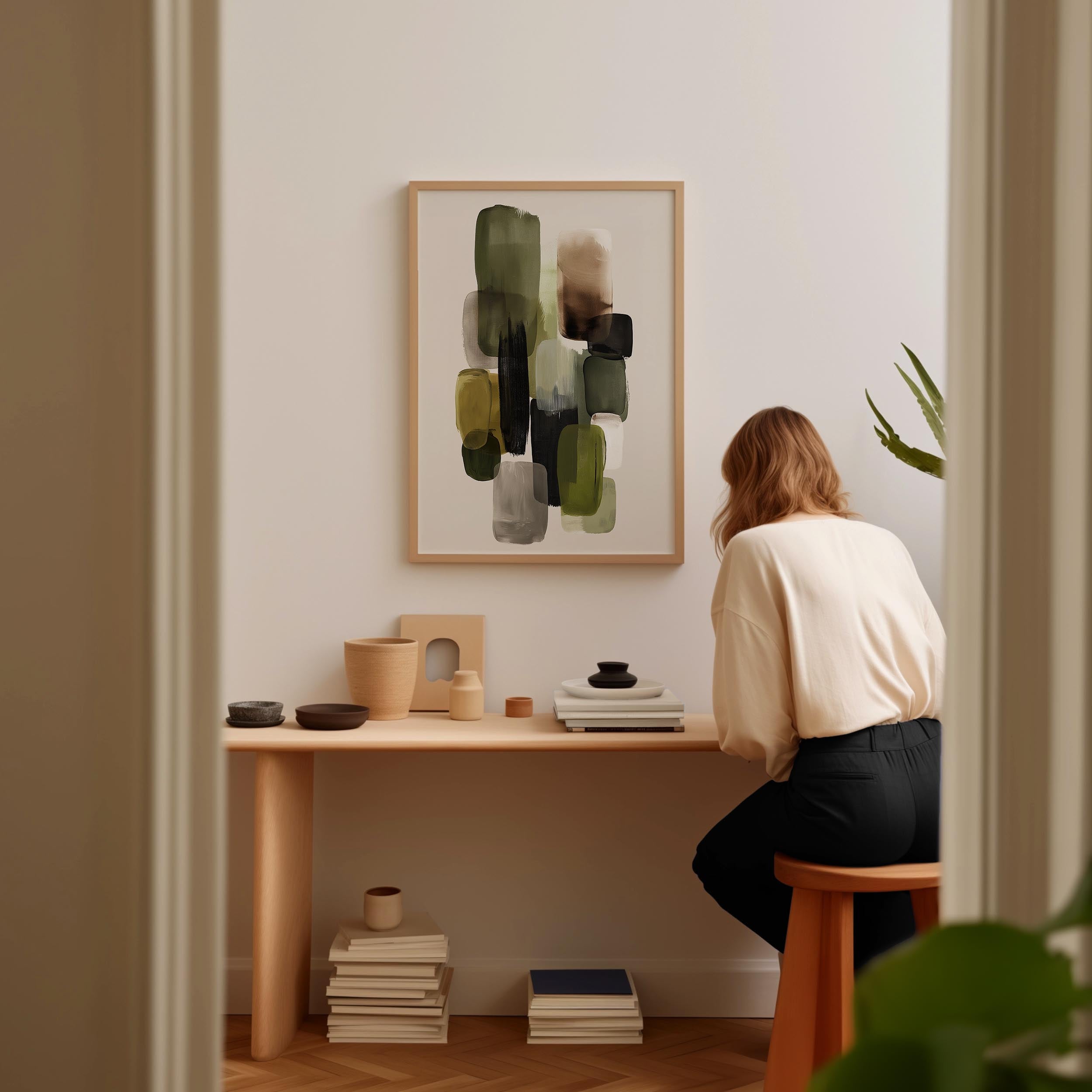 A woman is sitting at a wooden desk, surrounded by various objects, including a framed abstract painting, a stack of books, and a plant.