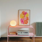 a pink metal table with a framed artwork of colorful flowers on it, a lamp, and a radio on the lower shelf.