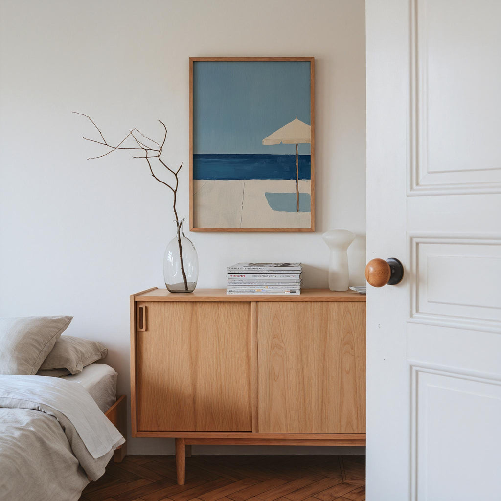 a serene and minimalist bedroom scene, featuring a wooden dresser with a framed painting of a beach scene above it, a vase with branches, and a stack of books on the dresser.