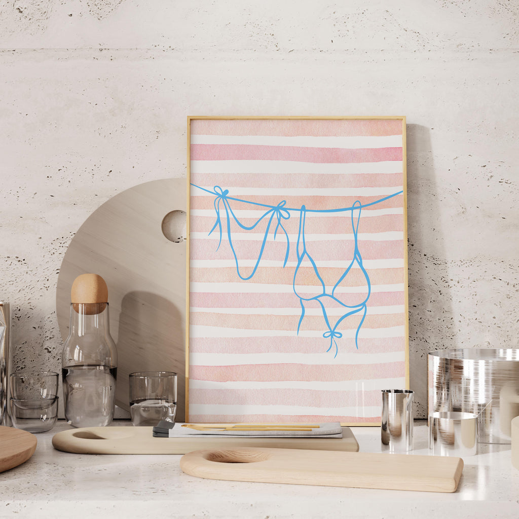 a framed artwork with a pink and white striped background, depicting a pair of blue underwear hanging on a clothesline. The artwork is displayed on a shelf, surrounded by various kitchen items such as a bottle, a glass, and a wooden cutting board.