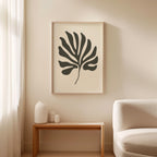 a minimalist living room with a large black leaf-shaped artwork hanging on the wall above a wooden side table. The table has three small white vases on it. The room has a white sofa and a wooden coffee table in front of it.