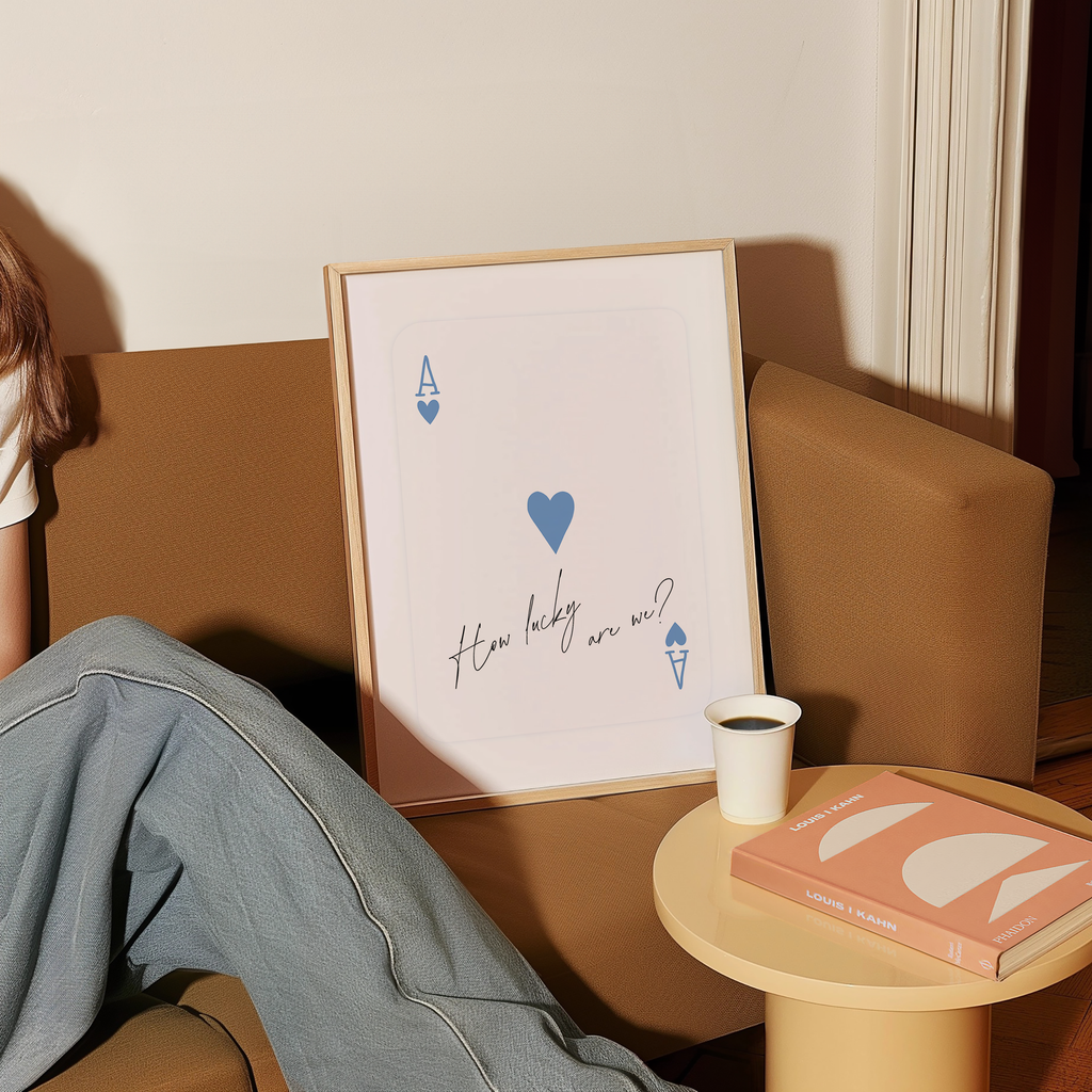 a person sitting on a couch with a framed poster of a playing card with a heart and the text "How lucky are we?" next to a small table with a book and a cup of coffee.