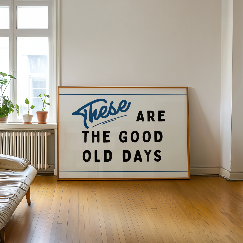 a large framed sign with the text "These are the good old days" on it, placed on a wooden floor in a room with a window and a plant.
