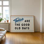 a large framed sign with the text "These are the good old days" on it, placed on a wooden floor in a room with a window and a plant.