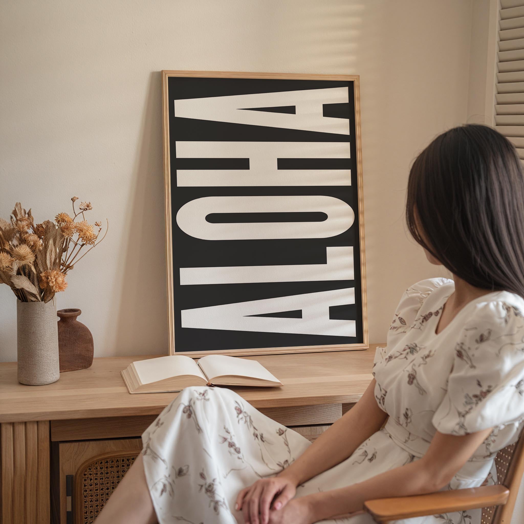 A woman in a white dress is sitting at a desk with an open book and a vase of flowers in front of her.