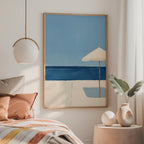 a serene bedroom scene with a large painting of a beach scene hanging on the wall, featuring a blue sky, ocean, and a white umbrella. The painting is framed in a wooden frame and is placed above a bed with a striped blanket. To the side of the bed, there is a small table with a vase and a plant, adding a touch of greenery to the room.
