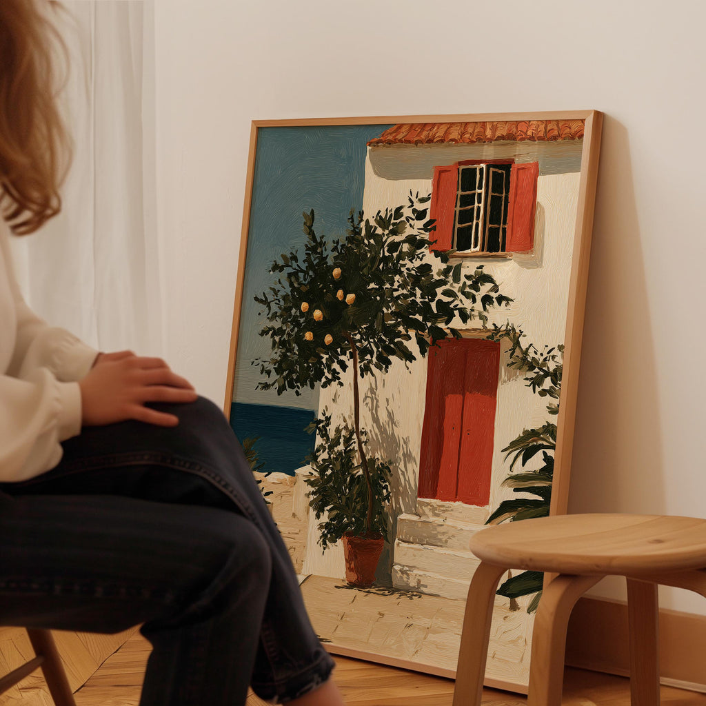 A person is sitting in a chair in front of a large painting depicting a white house with a red door and a tree in front of it.