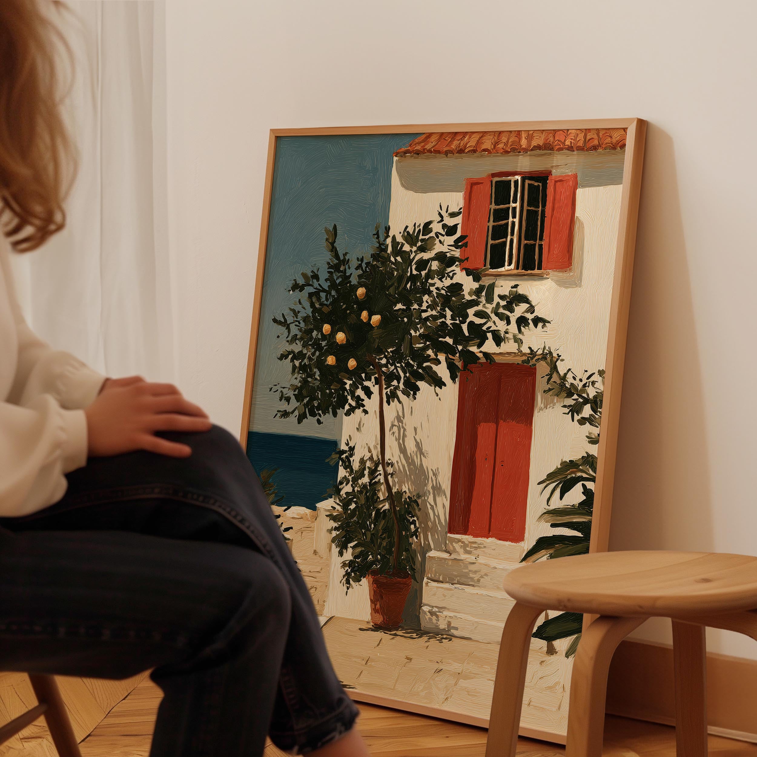 A person is sitting in a chair in front of a large painting depicting a white house with a red door and a tree in front of it.
