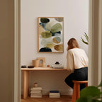 A woman is sitting at a wooden desk, surrounded by various objects, including a stack of books, a plant, and a framed abstract art piece on the wall.