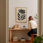 A woman is sitting at a wooden desk, surrounded by various objects, including a framed plant-like artwork, books, and a stack of books.
