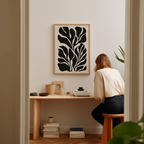 A woman is sitting at a wooden desk, surrounded by various objects, including a stack of books, a plant, and a framed abstract art piece on the wall.