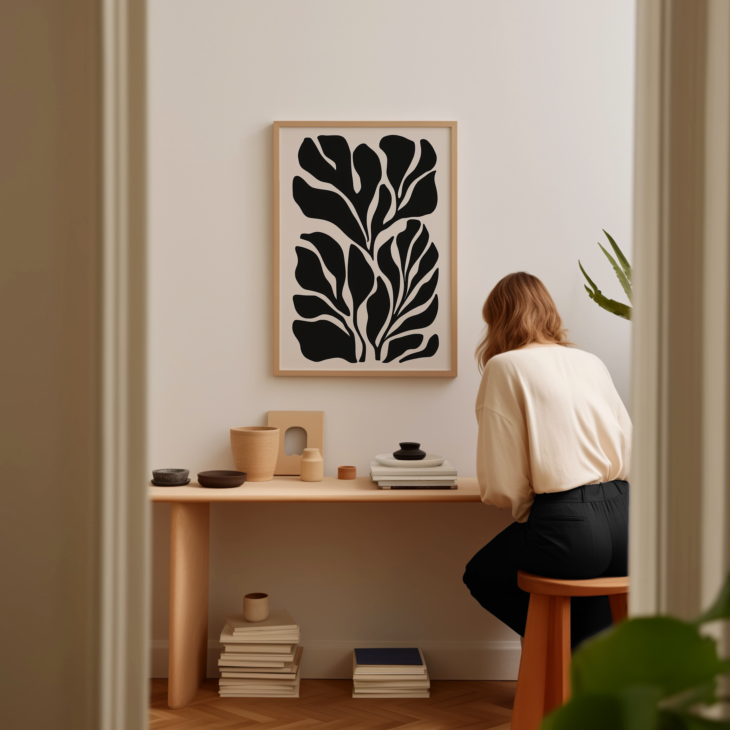 A woman is sitting at a wooden desk, surrounded by various objects, including a stack of books, a plant, and a framed abstract art piece on the wall.