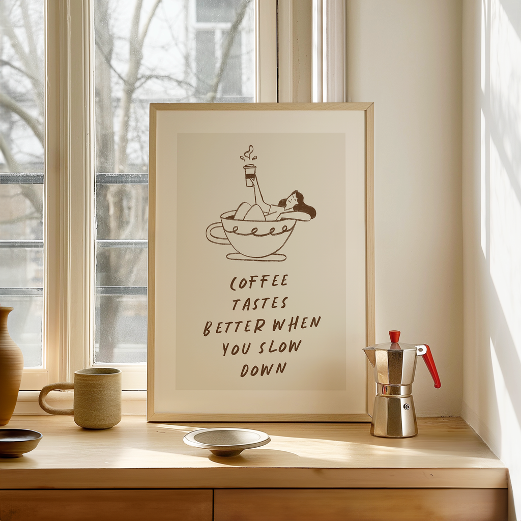 a framed poster with a cartoon illustration of a woman in a cup of coffee, accompanied by the text "Coffee tastes better when you slow down". The poster is displayed on a wooden shelf, with a coffee maker and other decorative items visible in the background.