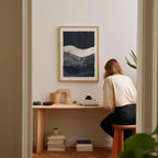 A woman is sitting at a wooden desk, surrounded by various objects, including a framed abstract painting, a stack of books, and a plant.