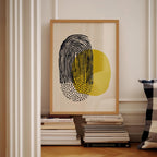 a framed abstract art piece with a black and yellow design, placed on a shelf next to a stack of books.