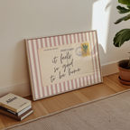 A framed postcard with a handwritten message sits on a wooden floor next to a stack of books and a potted plant.