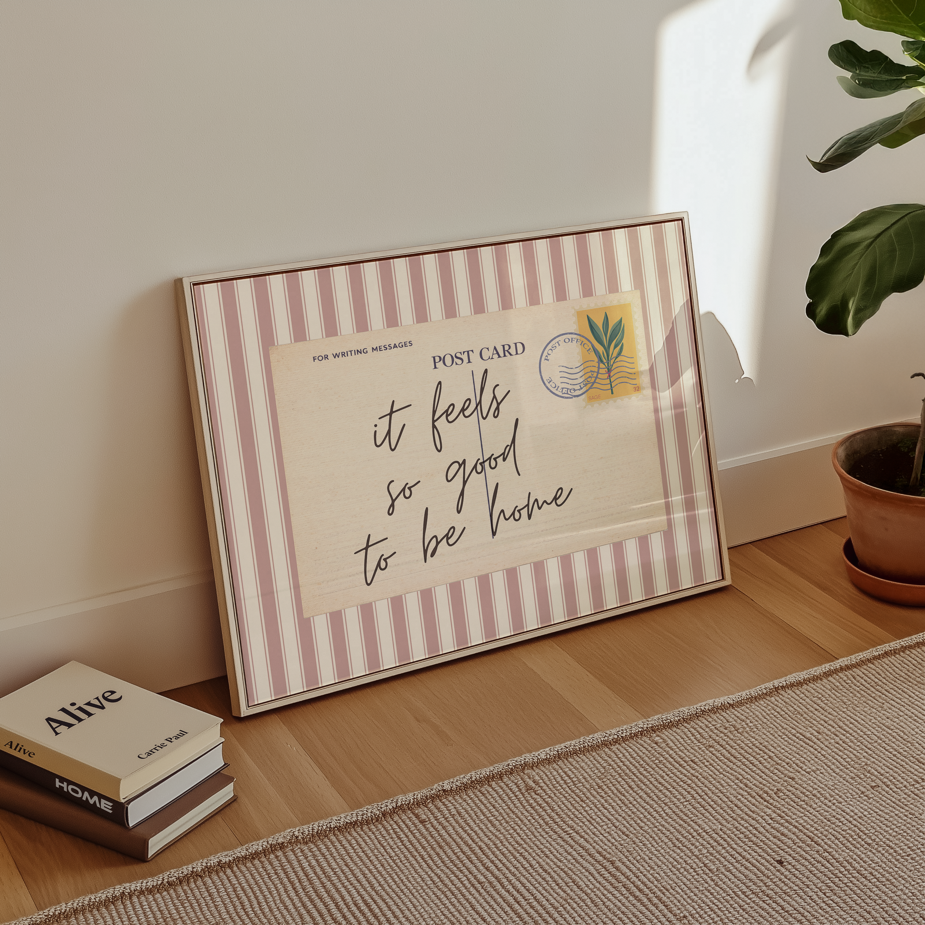 A framed postcard with a handwritten message sits on a wooden floor next to a stack of books and a potted plant.
