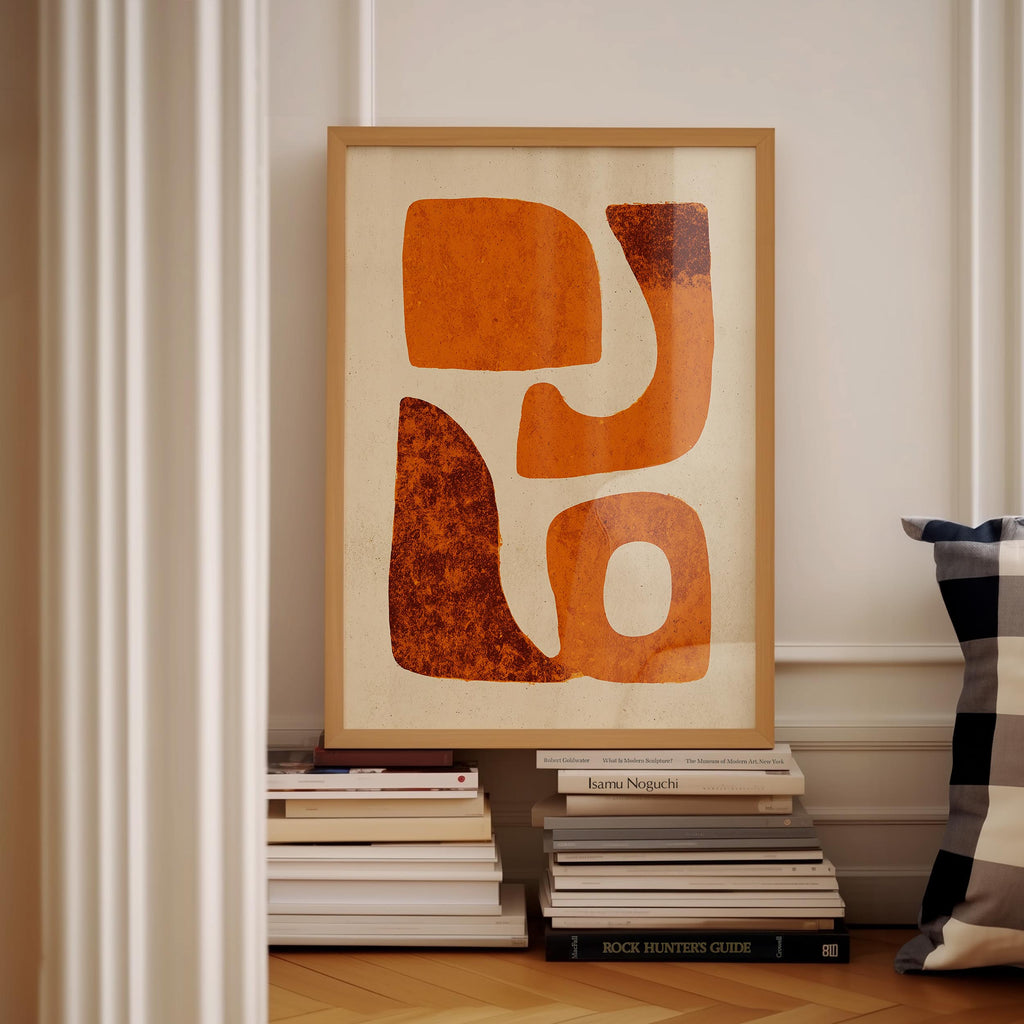 a framed abstract artwork with orange and brown shapes, displayed on a shelf among a stack of books.