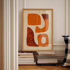 a framed abstract artwork with orange and brown shapes, displayed on a shelf among a stack of books.