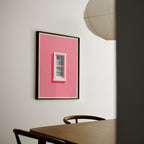 a pink wall with a framed artwork depicting a window, and a wooden table with two chairs in the foreground.