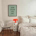 a bedroom with a bed, a nightstand, and a chair. The bed has white bedding and a green and white striped poster on the wall. The nightstand has a red lamp on top. The chair is empty.