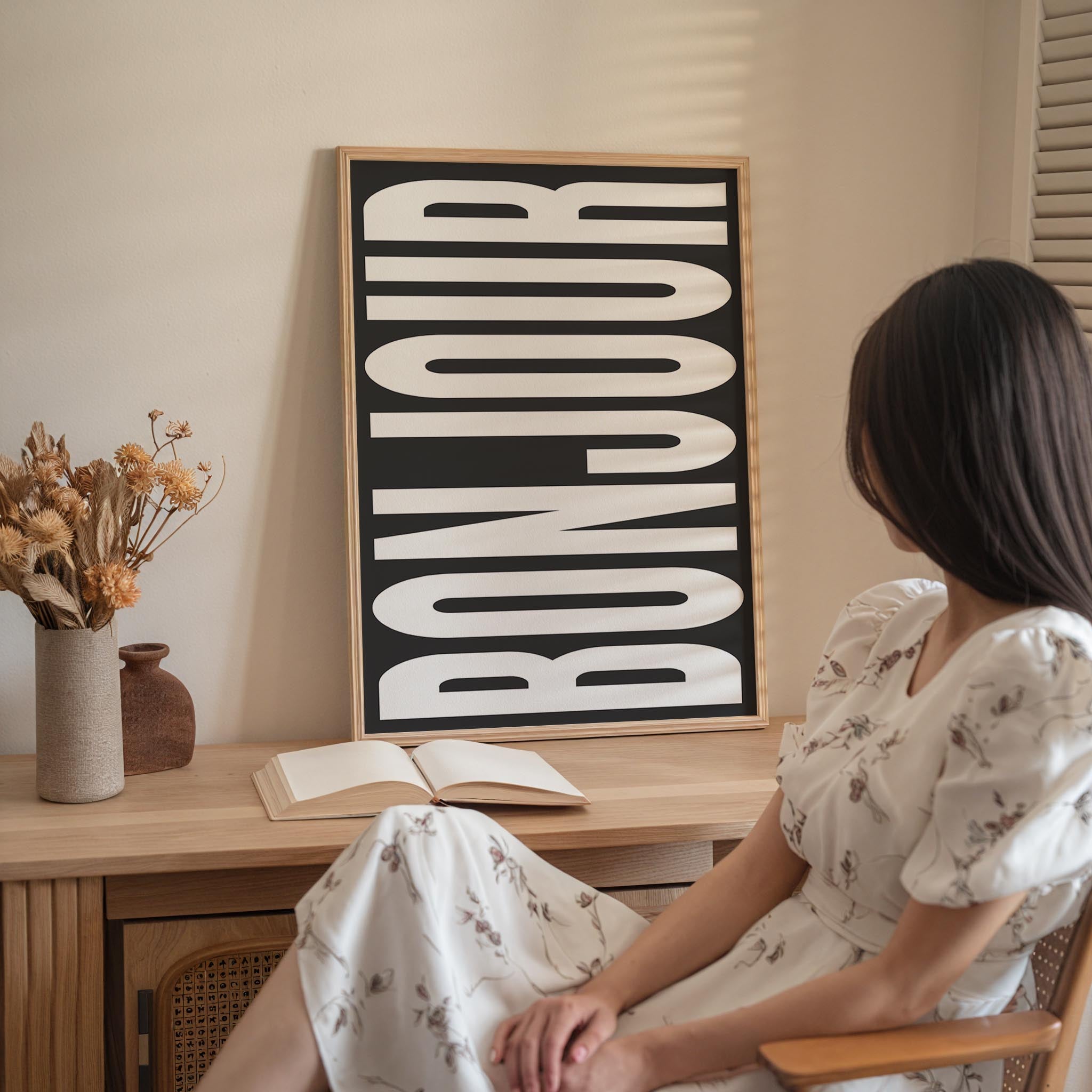 A woman in a white dress is sitting at a desk with an open book and a vase of flowers, with a large framed sign that says " bonjour" in the background.