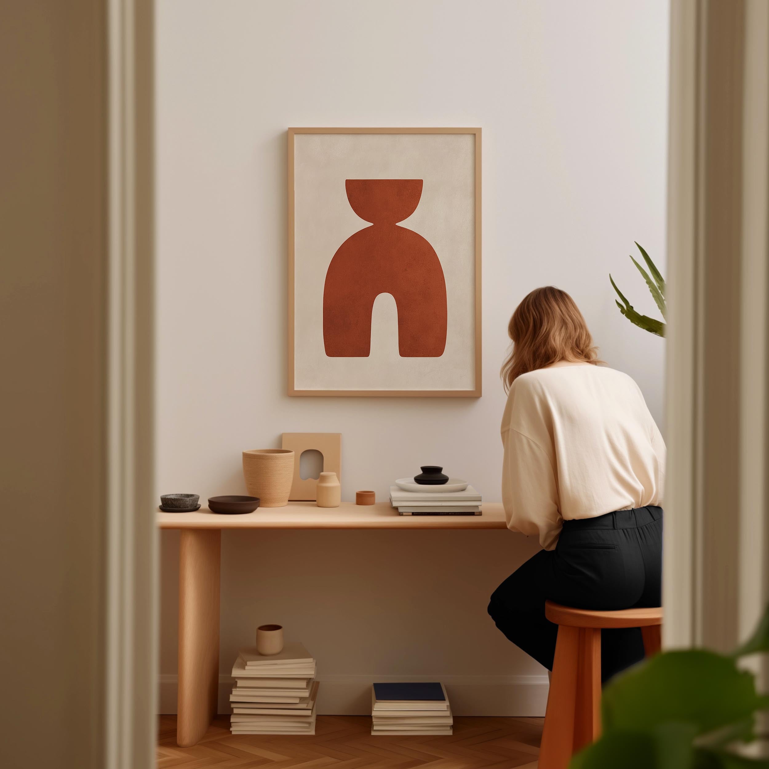 A woman is sitting at a wooden desk, surrounded by various objects, including a framed abstract artwork, a stack of books, and a plant.