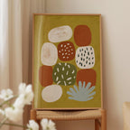 a framed abstract art piece with various shapes and colors, including white, orange, green, and brown, displayed on a wooden stand.