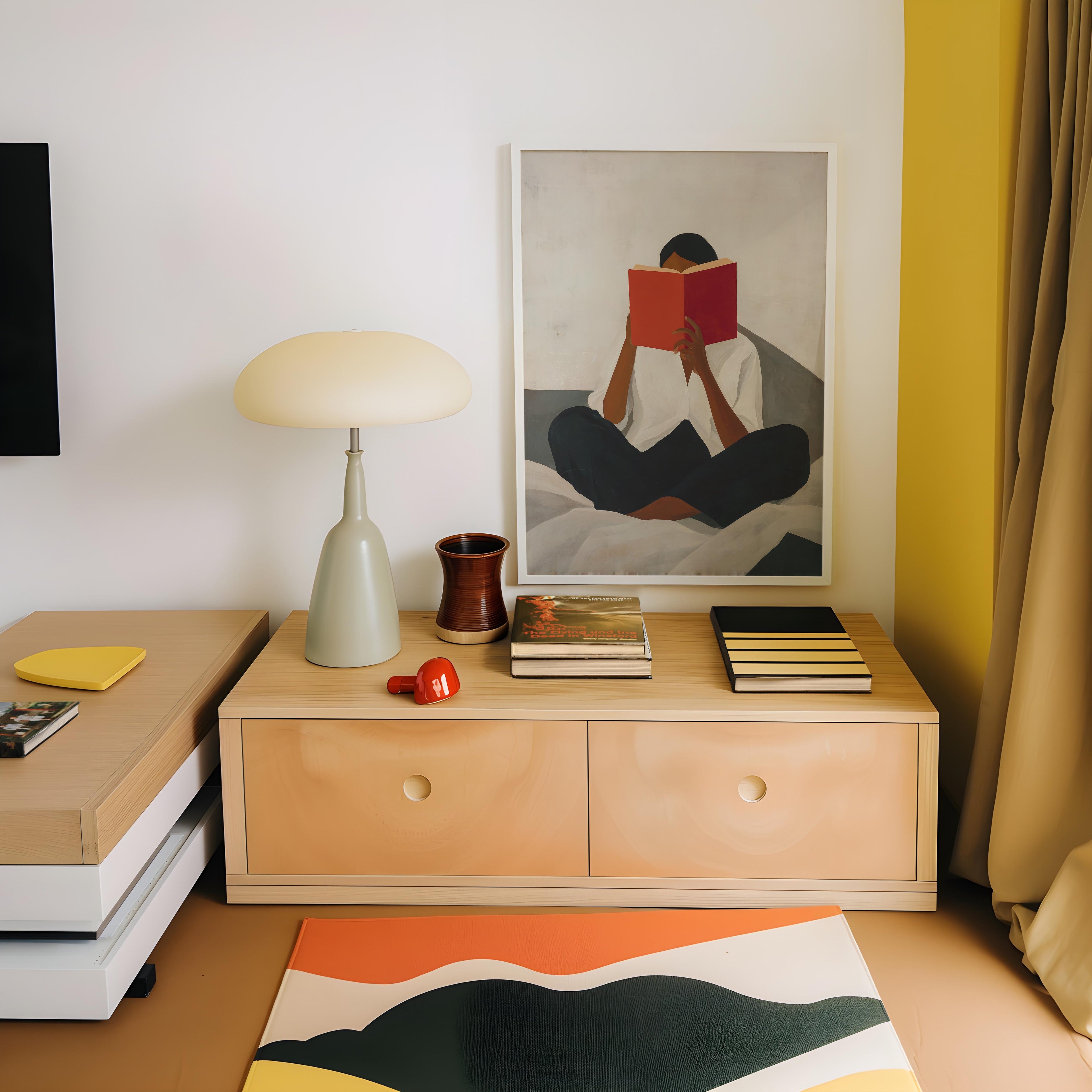 a cozy corner of a room with a wooden dresser, a lamp, and a framed painting of a person reading a book. The dresser has two drawers and a small table with a lamp on top. The painting depicts a person sitting on a bed and reading a book. The room has a warm and inviting atmosphere, with a beige wall and a yellow curtain.