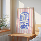 a framed poster of a can of Sardine with a decorative design, placed on a wooden table in a bedroom setting.