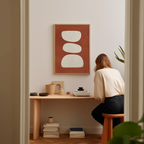 A woman is sitting at a wooden desk, surrounded by various objects, including a stack of books, a plant, and a framed abstract artwork on the wall.