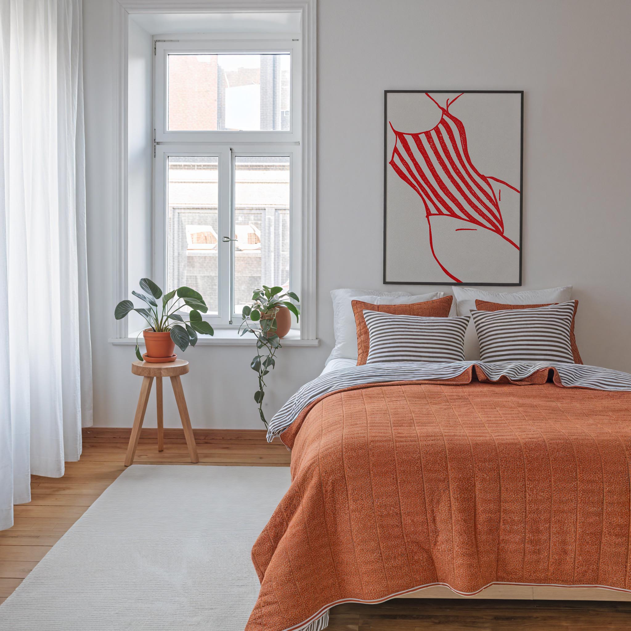 a cozy bedroom with a large bed, a wooden stool with a potted plant, and a large window with white curtains. The room has a warm, inviting atmosphere with the orange bedspread and striped pillows.