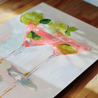 A painting of a pink cocktail glass with lime slices and green leaves, set against a wooden background.