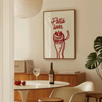 a dining room with a framed poster of a fork and pasta on the wall, a table with a wine bottle and wine glass, and a potted plant in the corner.
