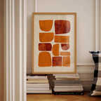 a framed abstract artwork with orange and brown shapes, displayed on a shelf among a stack of books.