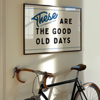 a framed sign with the text "These are the good old days" hanging on a wall next to a bicycle.