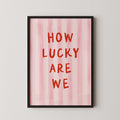 a framed poster with the text "HOW LUCKY ARE WE" written in bold red letters against a pink striped background.