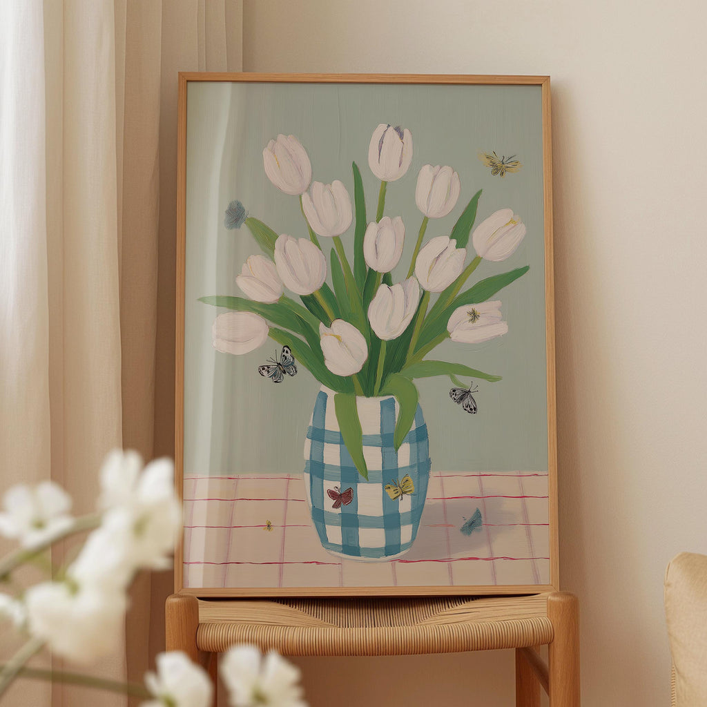 A framed painting of a vase filled with white tulips and butterflies is displayed on a wooden table.