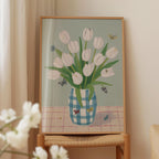 A framed painting of a vase filled with white tulips and butterflies is displayed on a wooden table.