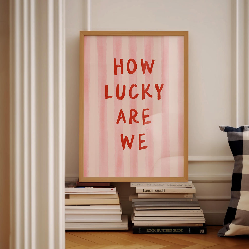 a framed poster with the text "HOW LUCKY ARE WE" displayed on it, placed on a shelf next to a stack of books.
