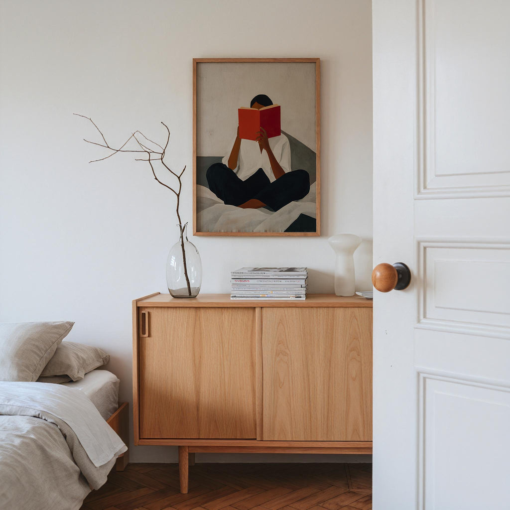 a cozy bedroom scene with a wooden dresser, a framed painting of a person reading a book, and a vase with branches.