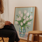 A person is sitting in front of a framed painting of a vase with white tulips and butterflies.