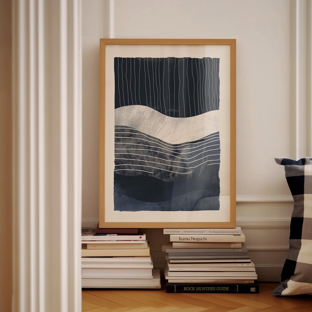 a framed abstract artwork with a dark blue and white color scheme, displayed on a white wall. Below the artwork, there is a stack of books and a pillow.