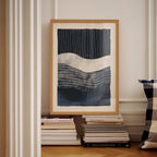 a framed abstract artwork with a dark blue and white color scheme, displayed on a white wall. Below the artwork, there is a stack of books and a pillow.