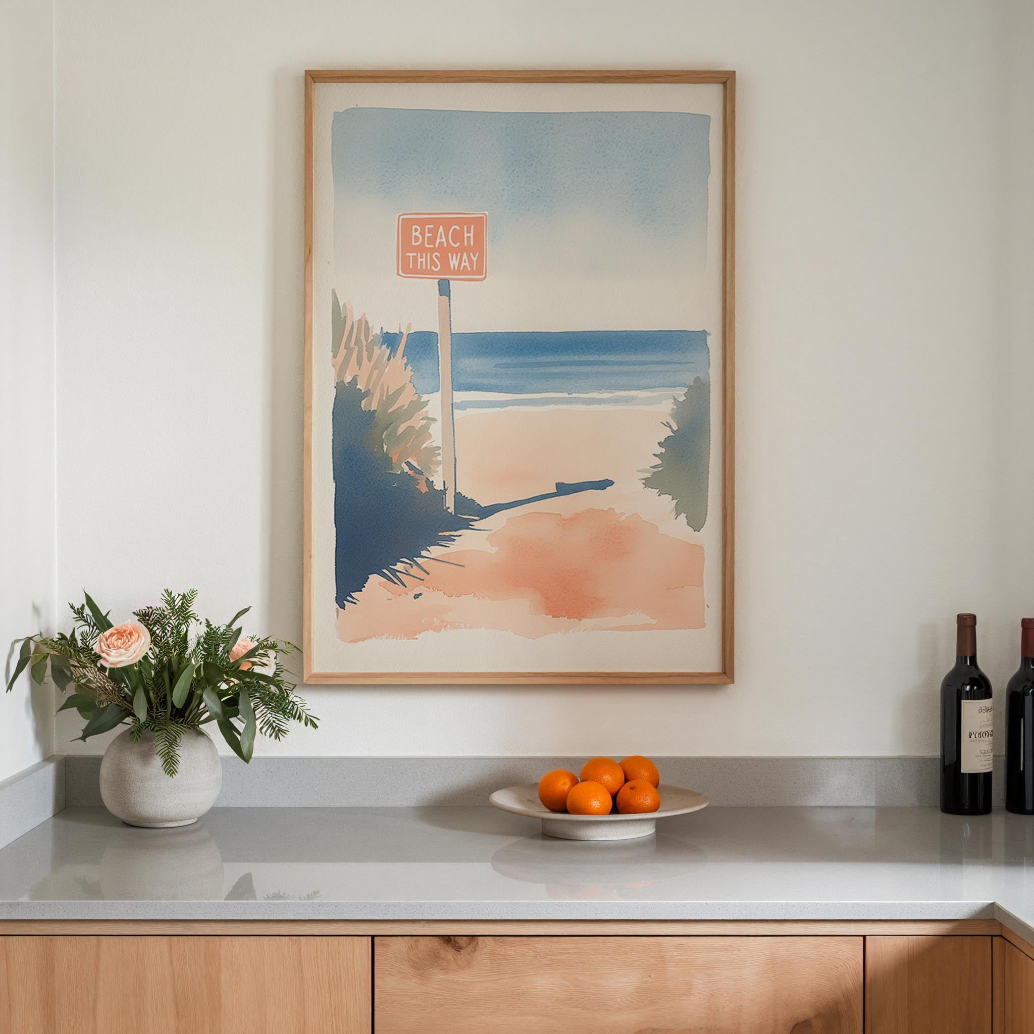 a framed painting of a beach scene with a sign that reads "BEACH THIS WAY", a vase of flowers, a plate of oranges, and two bottles of wine on a countertop.
