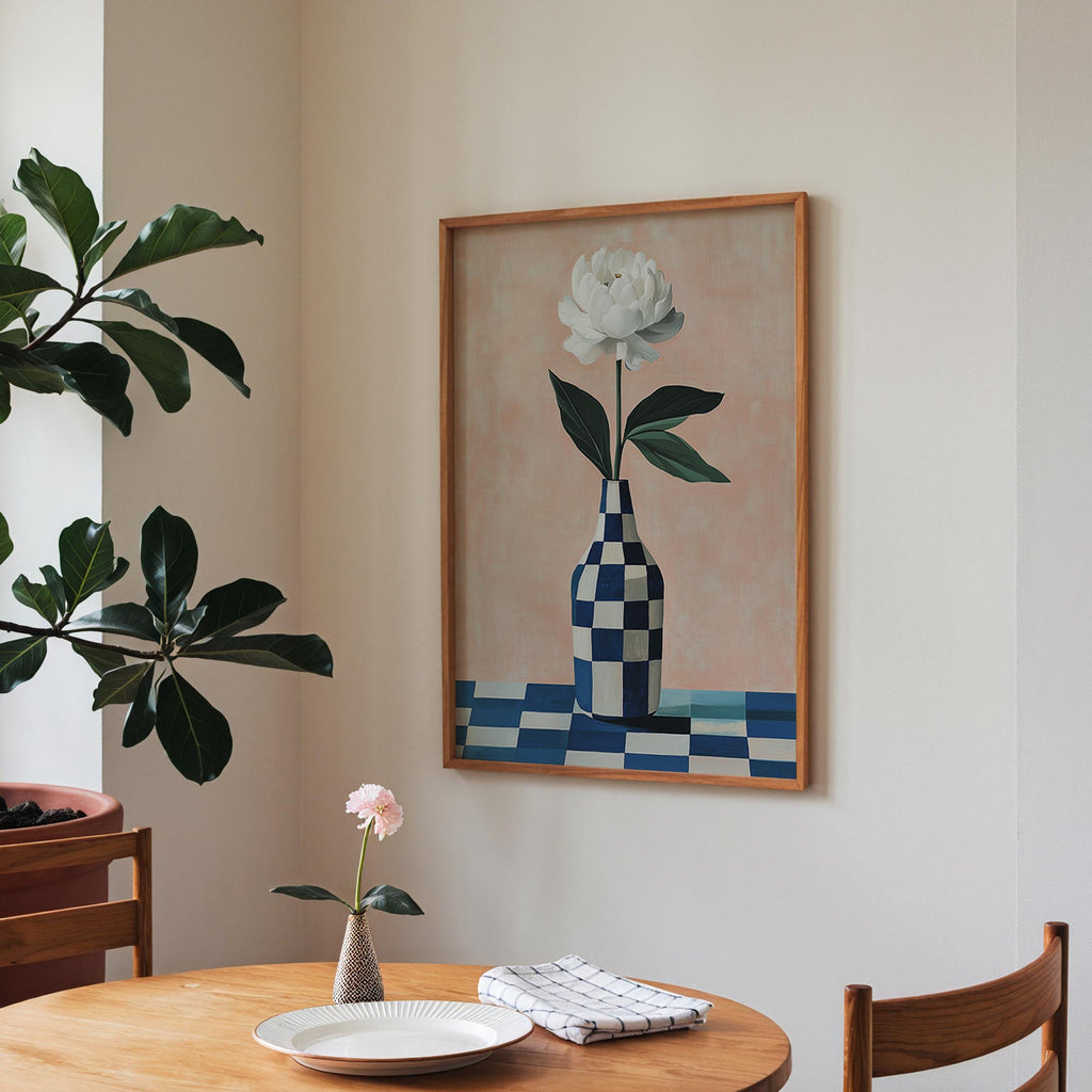 a dining area with a wooden table, a vase with a single pink flower, and a framed painting of a white flower in a blue and white checkered vase hanging on the wall.